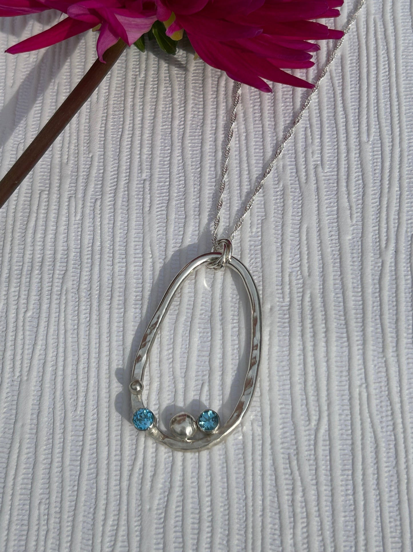 Handmade sterling silver pendant formed in a hammered oval shape with two blue topaz stones and silver nuggets. Hanging on a silver chain.